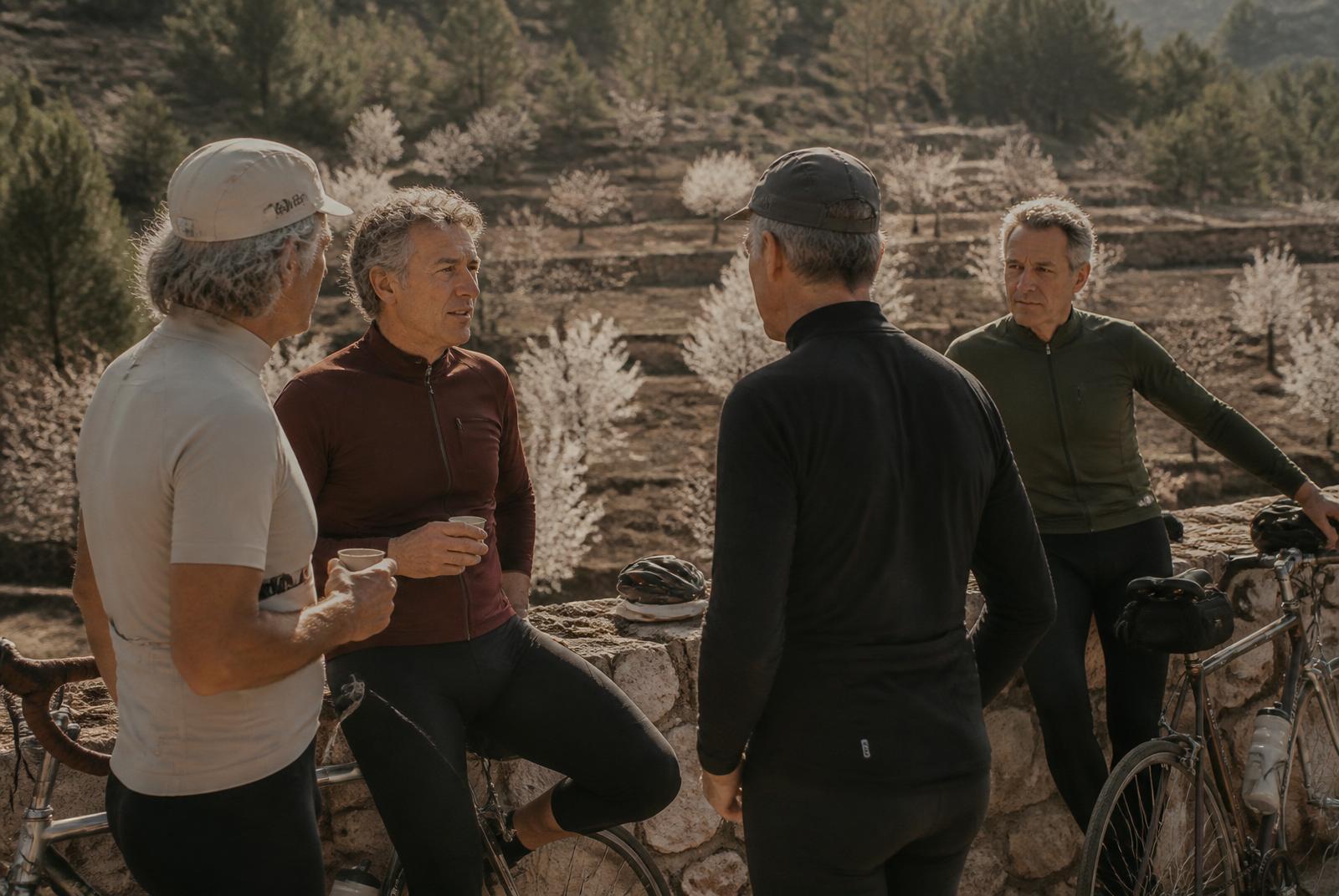 Pequeño grupo de ciclistas amateurs entre 55 y 65 años conversando junto a sus bicis frente a los almendros en flor de la Marina Alta