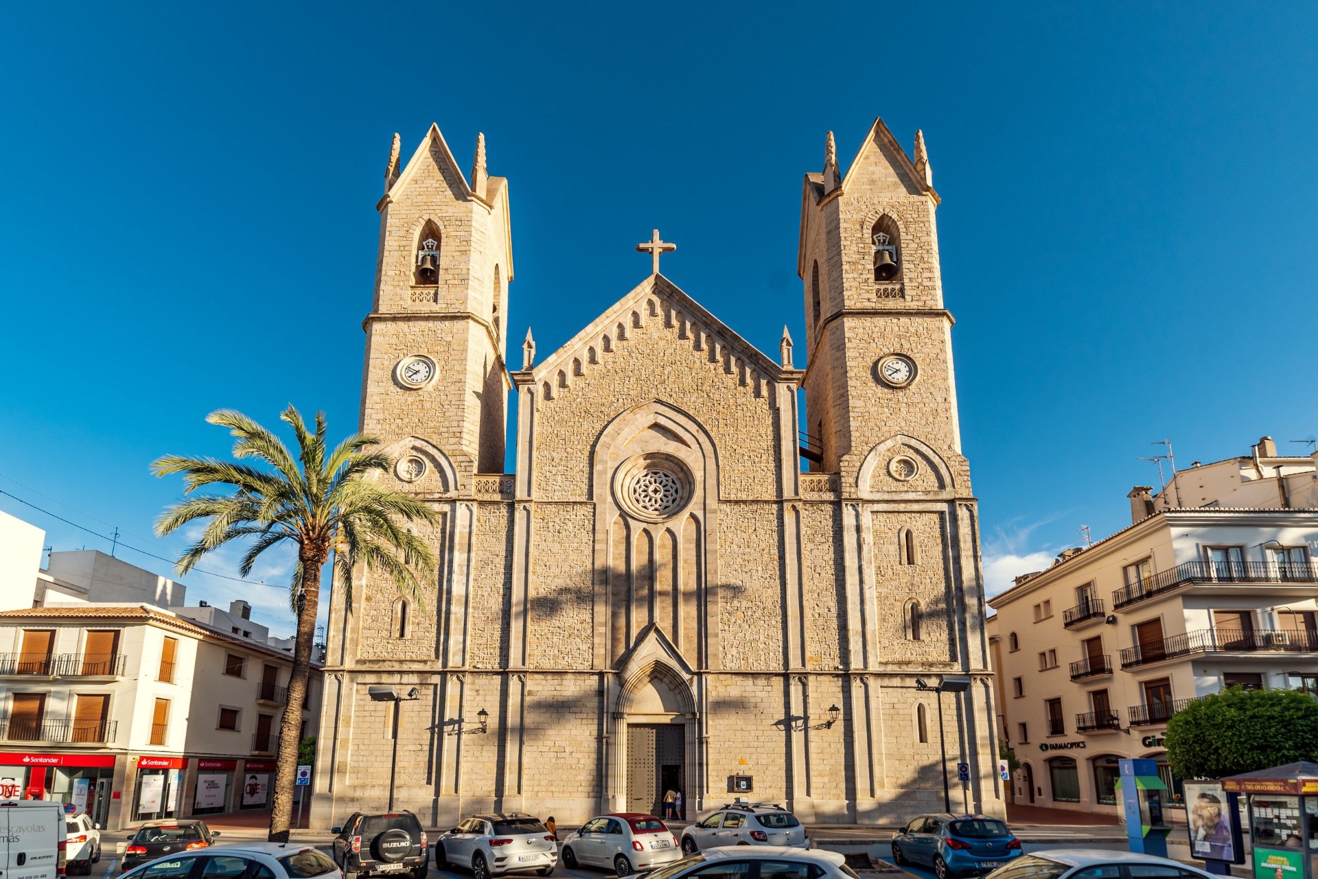 Fachada real de la Basílica Menor de la Puríssima Xiqueta i Sant Pere Apòstol en Benissa: piedra clara, dos campanarios simétricos con relojes, gran rosetón central y palmera en primer plano bajo cielo azul